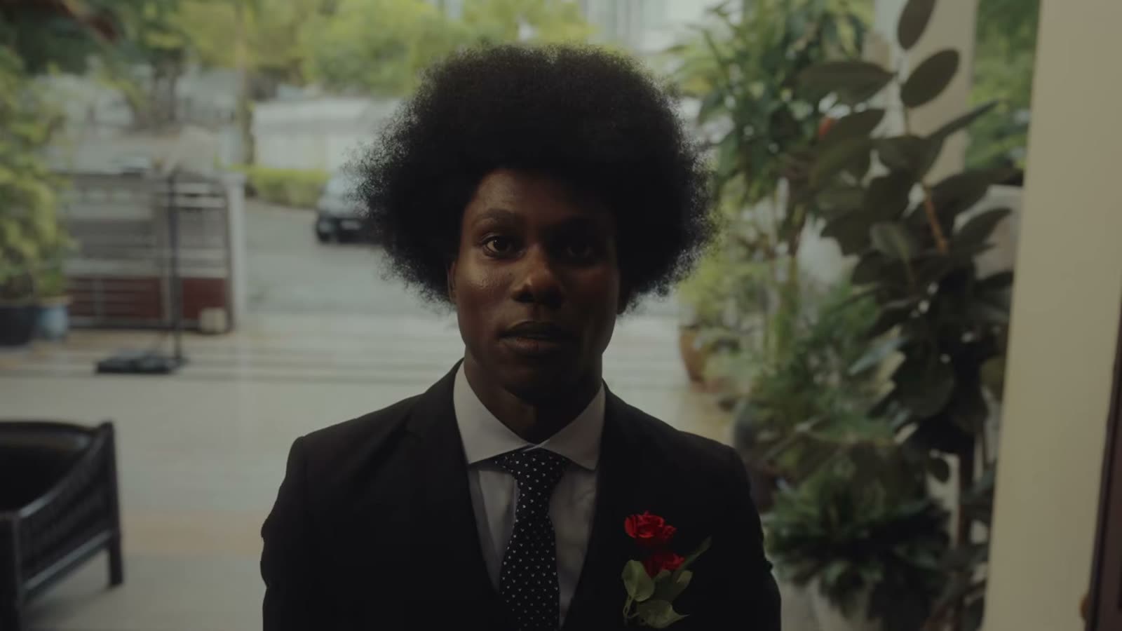 A young man with an afro, in a suit with a red flower on the lapel, waiting at a doorway