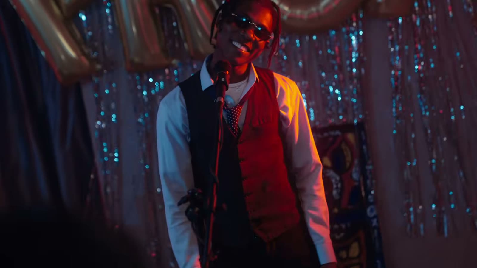 A performer in a waistcoat and sunglasses on a small stage under red light, with gold PROM balloons behind him